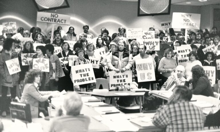 Large group of picketers holding signs supporting the New Bedford strike, standing behind a table filled with seated School Committee members.