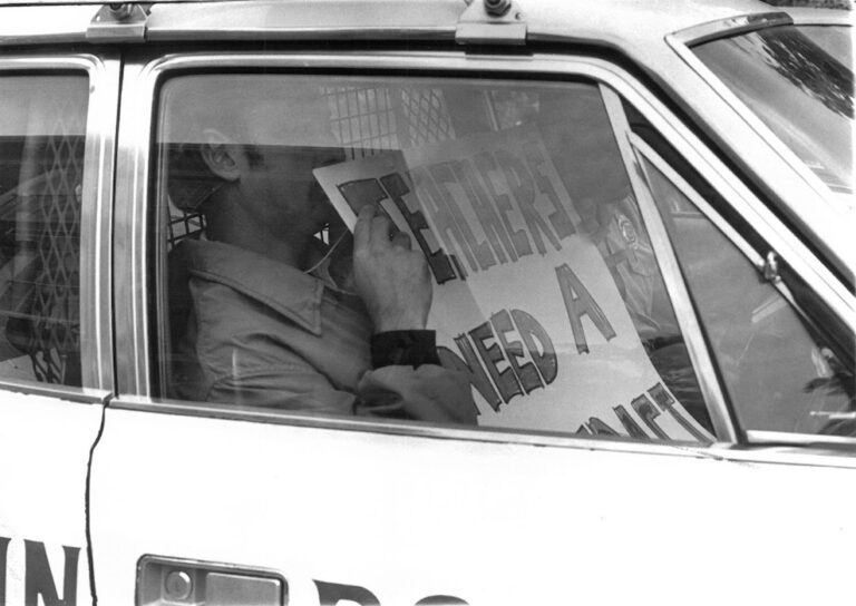 Picketer with sign in a police car.
