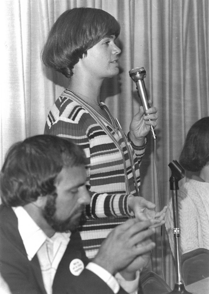 Person with a microphone standing and speaking.