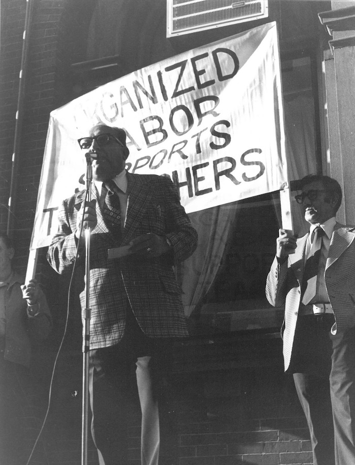 Man standing in front of microphone and behind a two people holding a sign.
