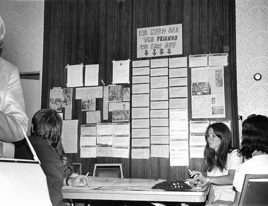 Group of people sitting in front of a wall covered in newspaper articles and other clippings.