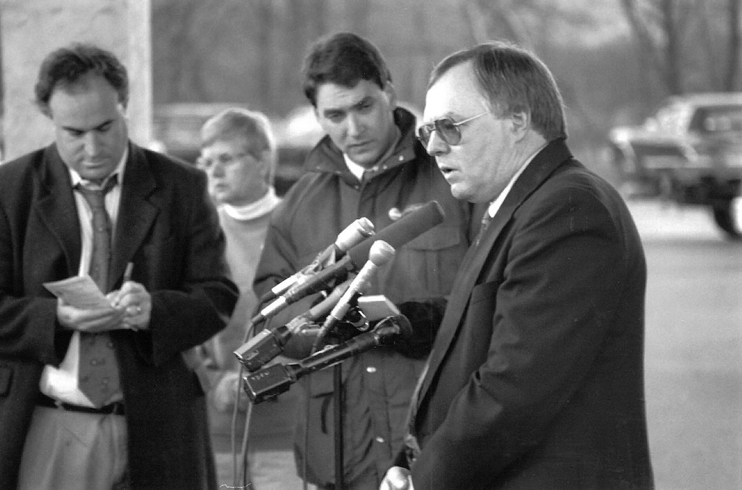 Side profile of person standing in front of microphones with people behind him.