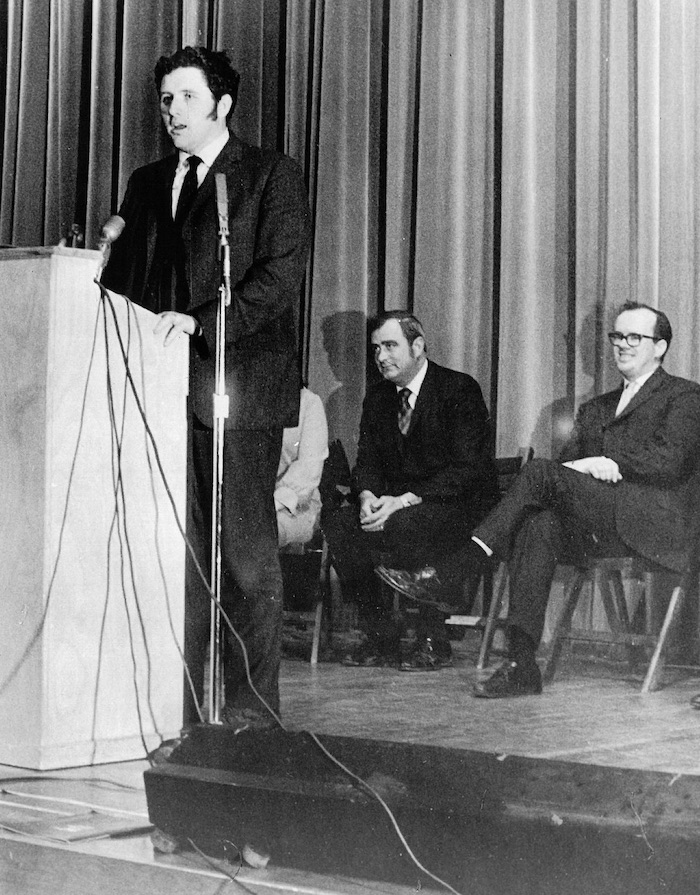 Person standing behind a podium with two people seated behind him.